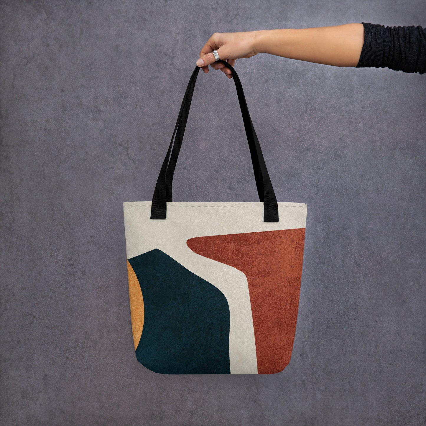 Person holding a HFT Tote Bag IV with abstract geometric design, showcasing rich earth tones setup against a textured gray background.